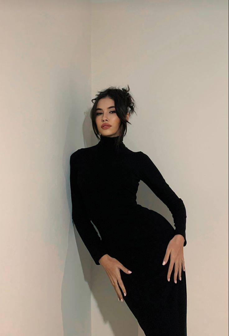 a woman leaning against a wall in a black dress with her hands on her hips