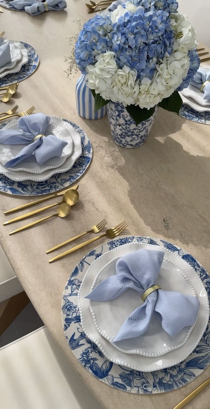 a table set with blue and white plates, gold place settings and flowers in vases