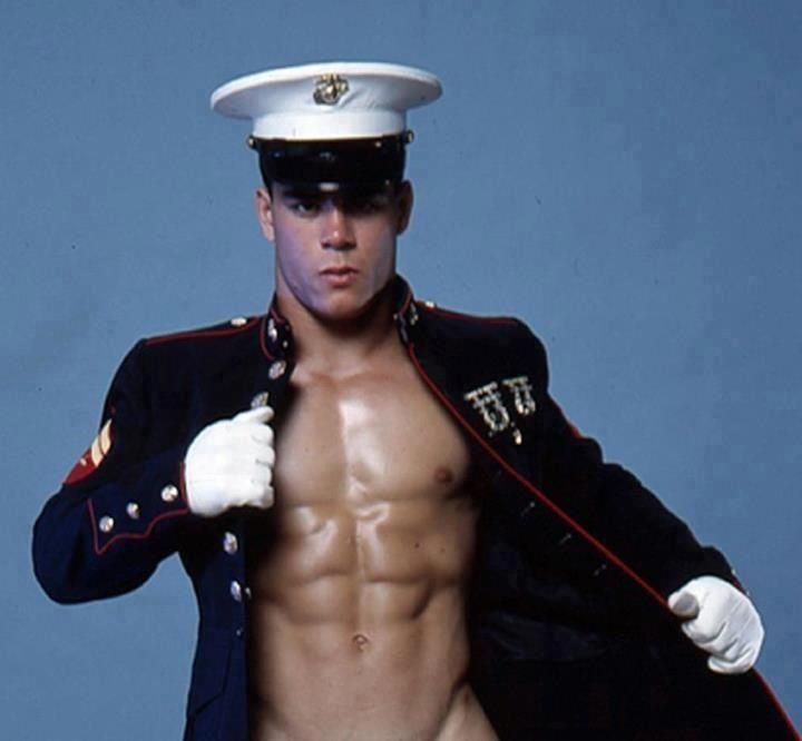 a shirtless man wearing a navy uniform and holding his jacket over his shoulder with one hand