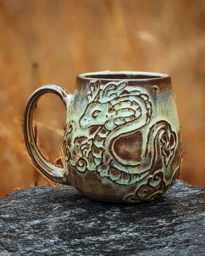 a ceramic mug with a dragon design on it sitting on a rock in front of some dry grass