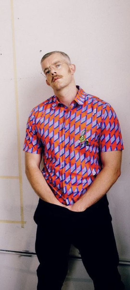 a man sitting on the floor with his hands in his pockets, wearing a colorful shirt and black pants