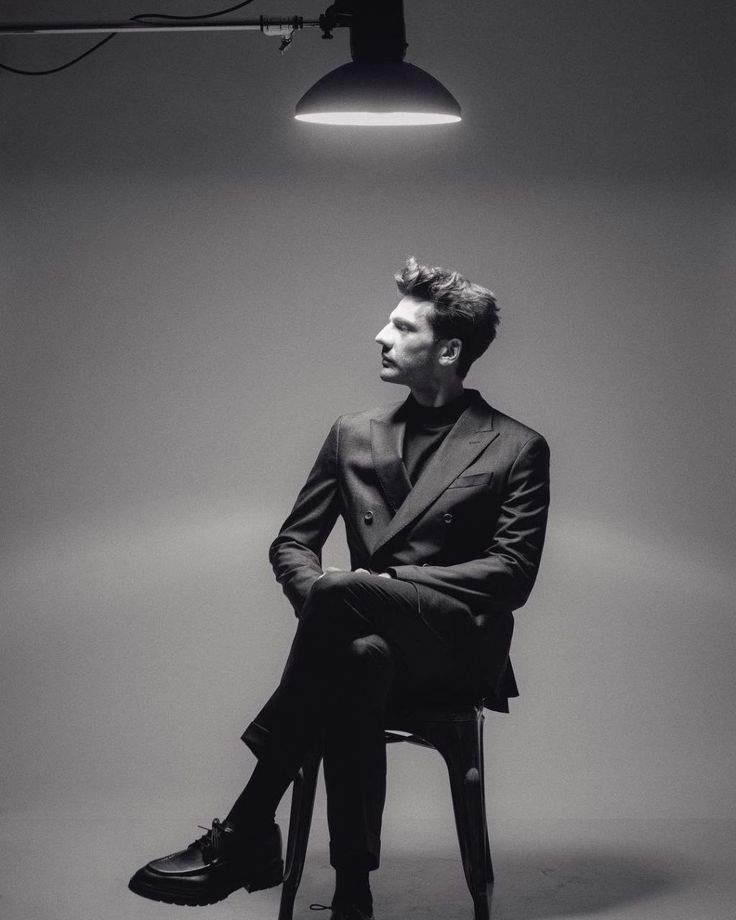 a man sitting on top of a chair in front of a light fixture and wearing a suit