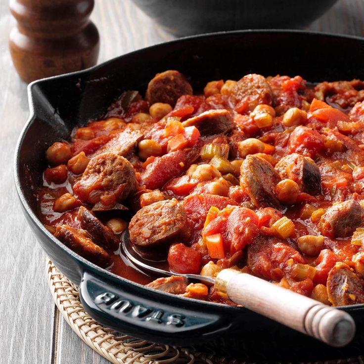 a pan filled with meat and beans on top of a wooden table next to a spoon