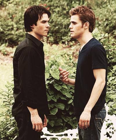 two young men standing next to each other in front of trees and bushes, talking