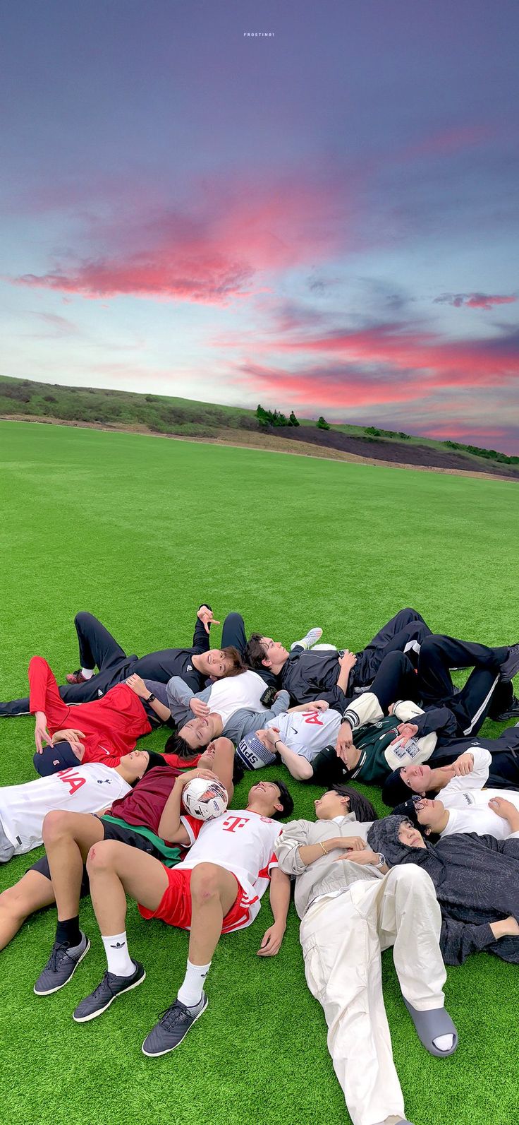 a group of people laying on top of a lush green field under a pink sky