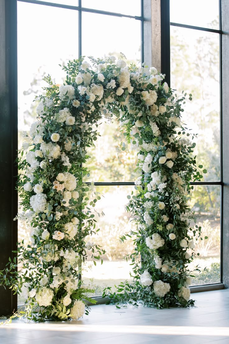 a floral arch with white flowers and greenery in front of a large glass window