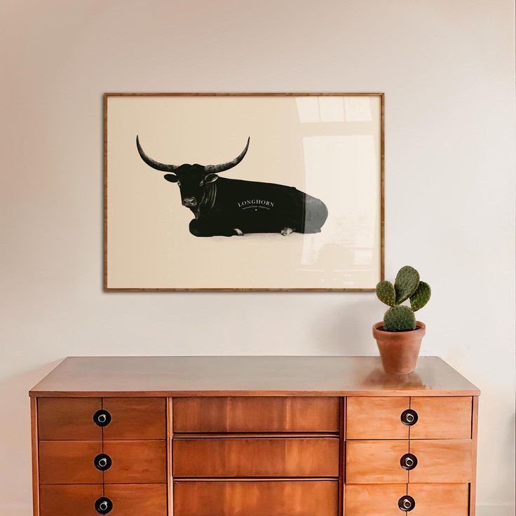 a cow is hanging on the wall above a dresser with a potted cactus in it