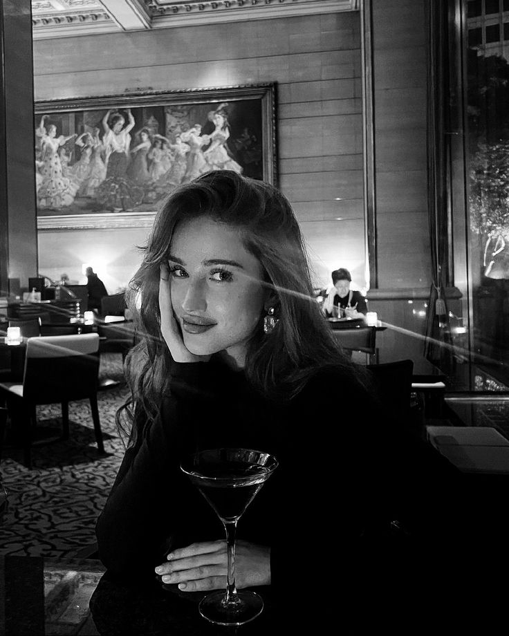 a woman sitting at a table with a wine glass in front of her and looking into the camera