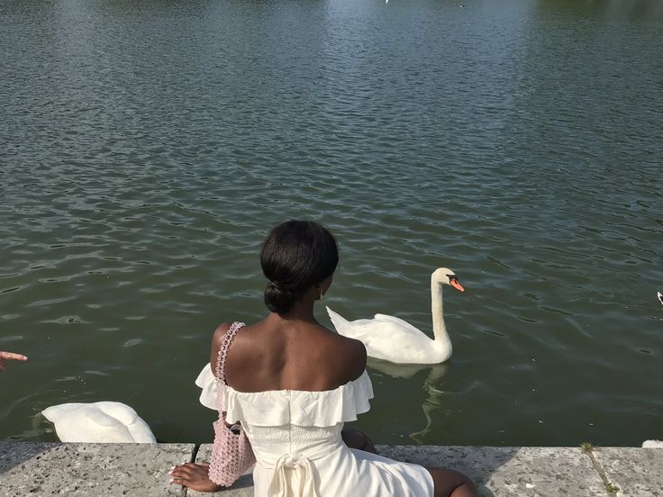 a woman sitting on the edge of a body of water with a swan in it