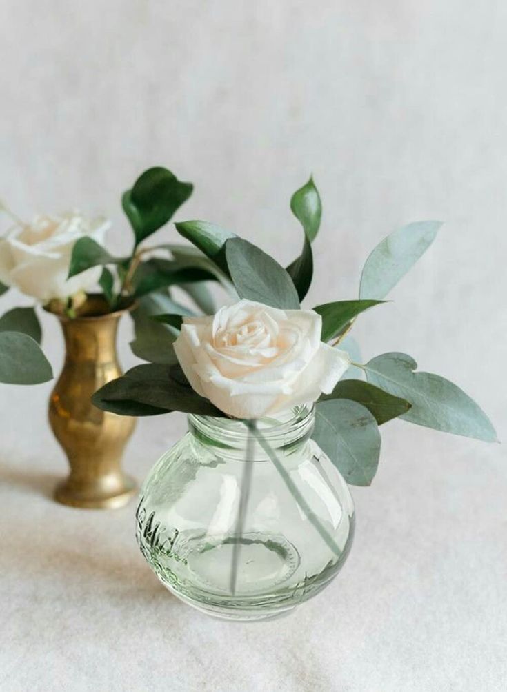 two small vases with flowers in them on a white tableclothed surface, one is gold and the other has green leaves