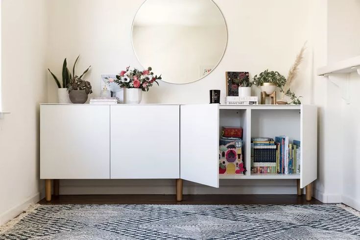 a white cabinet with books and plants on top next to a round mirror above it