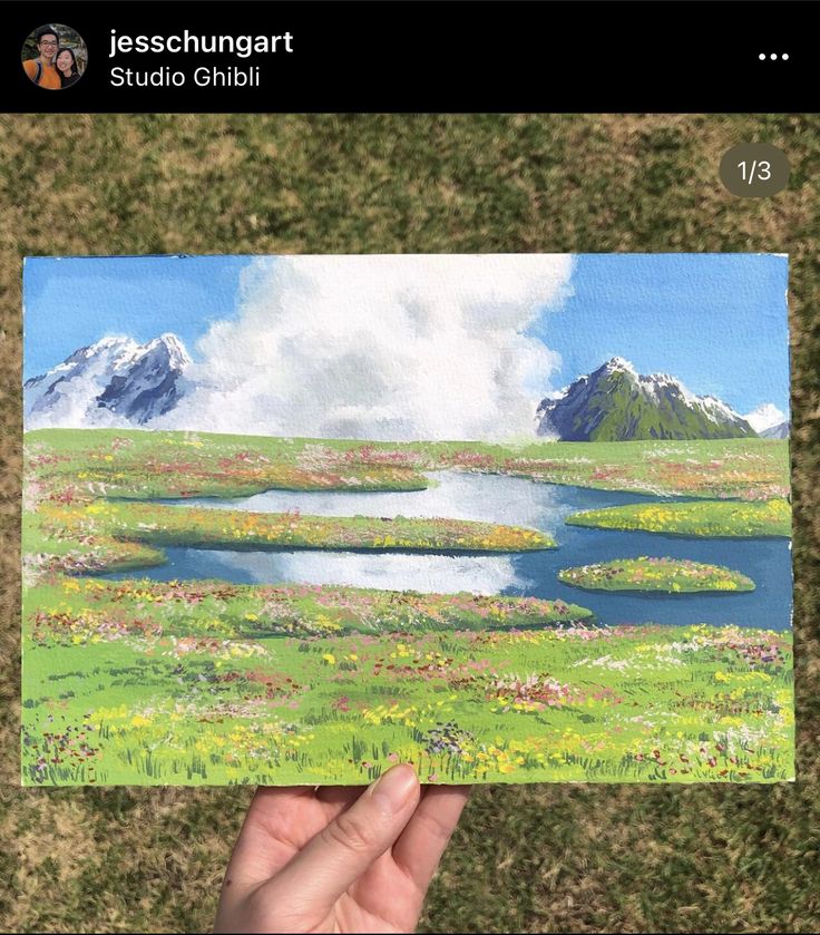 someone is holding up a painting in their hand, with mountains and flowers on the other side