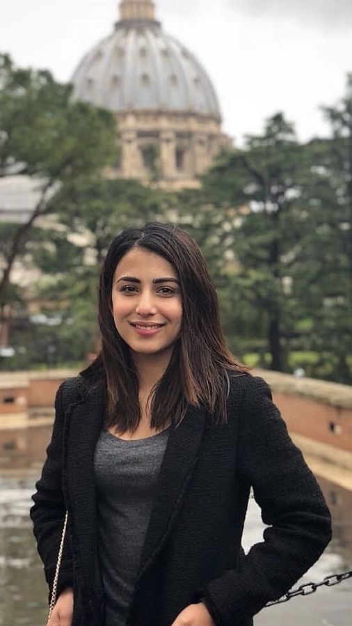 a woman standing in front of a fountain with a building in the back ground and trees behind her