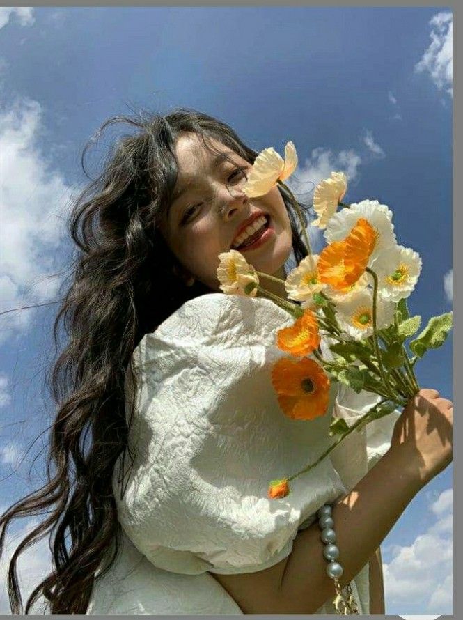 a woman holding flowers in her arms and smiling at the camera, against a blue sky with clouds