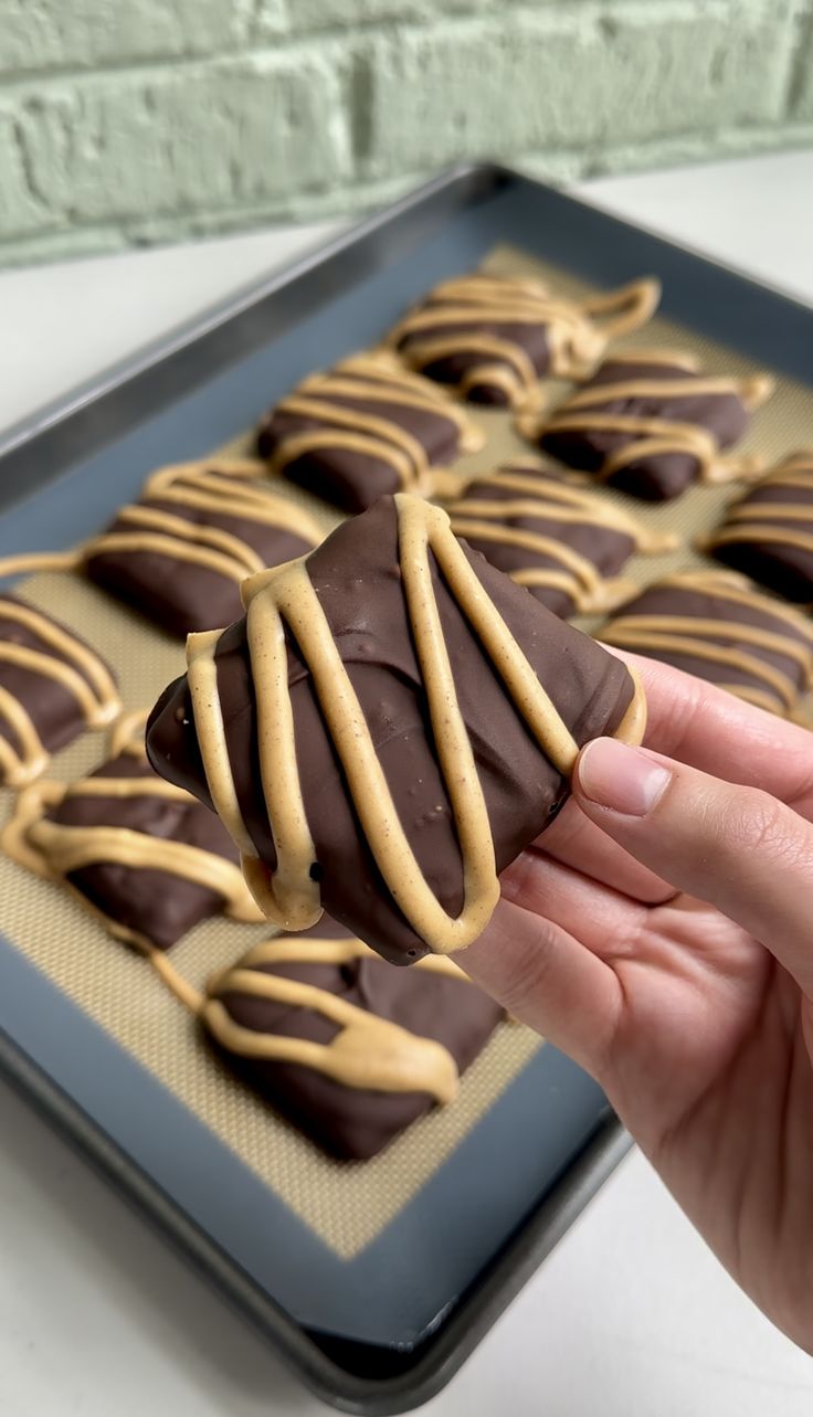 a hand holding a chocolate covered pretzel in front of a baking tray with more pretzels on it