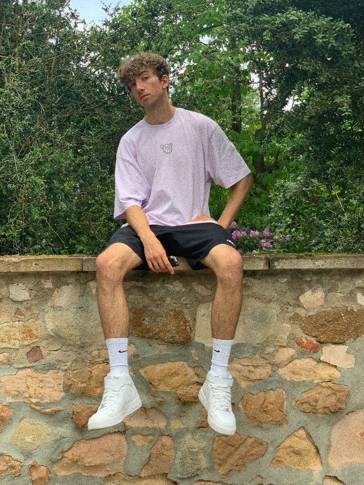 a man sitting on a stone wall with his legs crossed, wearing white sneakers and black shorts