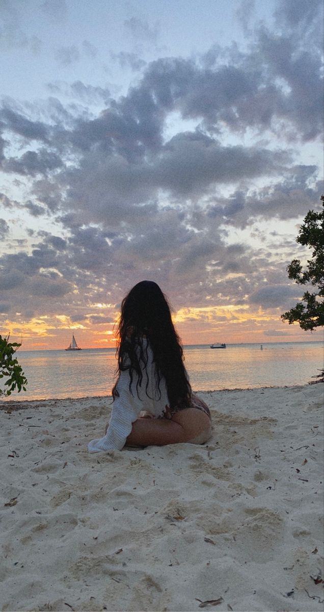 a woman is sitting on the beach at sunset