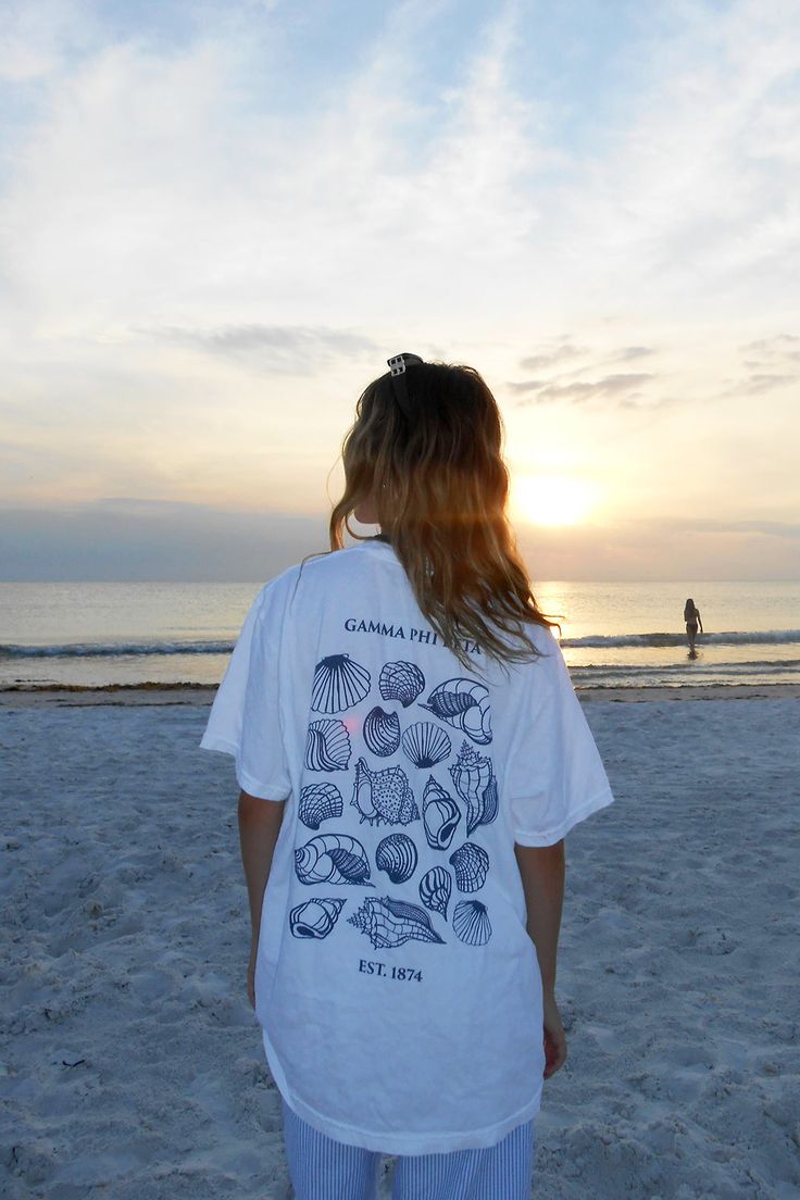 a woman standing on top of a sandy beach next to the ocean with shells printed on her t - shirt