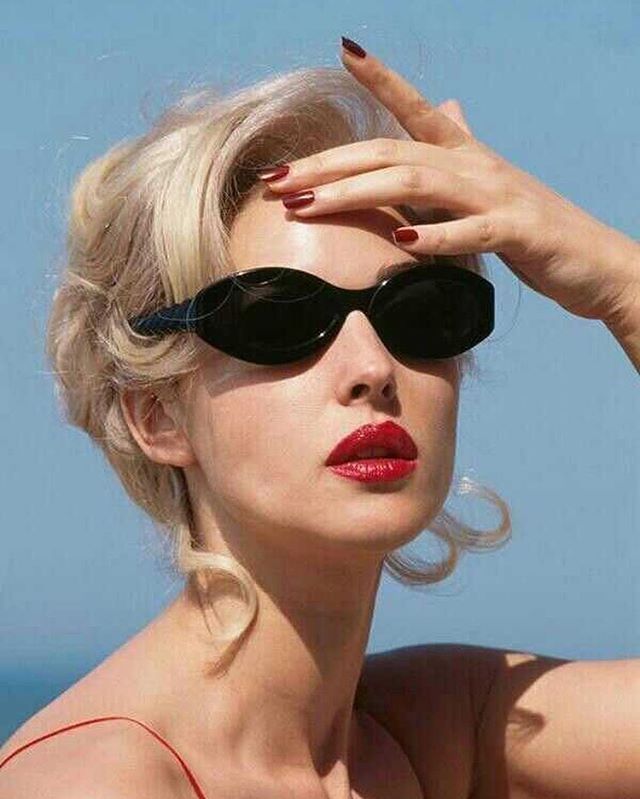 a woman with sunglasses on her head and red lipstick in front of the ocean, holding her hand up to her face