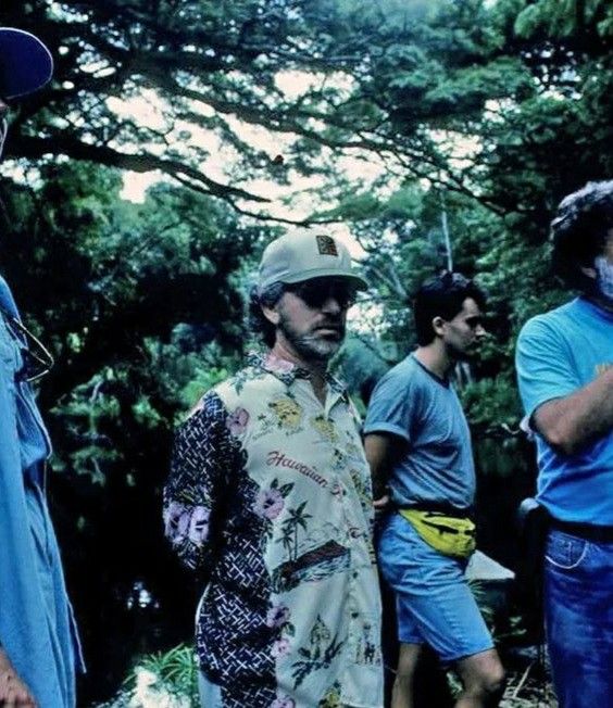 a group of men standing next to each other in front of trees and bushes with one man wearing a hat