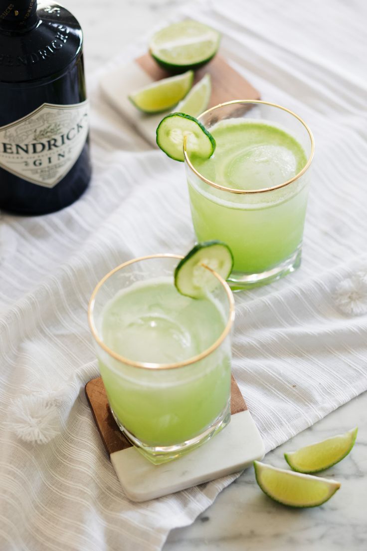 two glasses filled with green liquid and lime slices next to a bottle of gin tonic