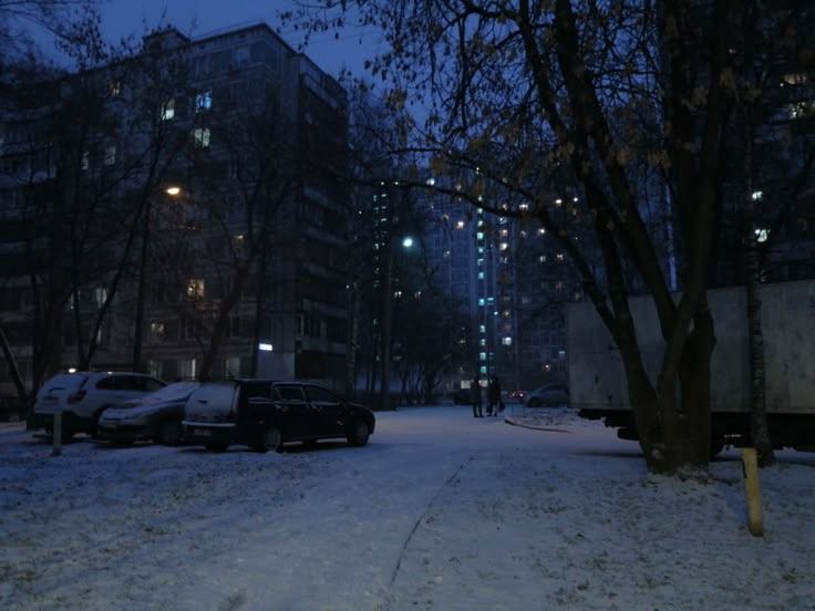 cars parked on the side of a snow covered road at night with buildings in the background