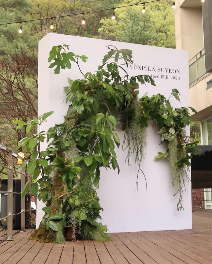 a sign with plants growing on it in front of a building that has trees and lights hanging from the roof