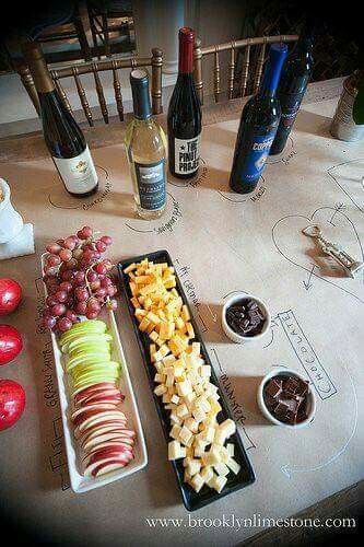 an assortment of fruits and cheeses on a table next to bottles of wine, chocolate sauce, and apples