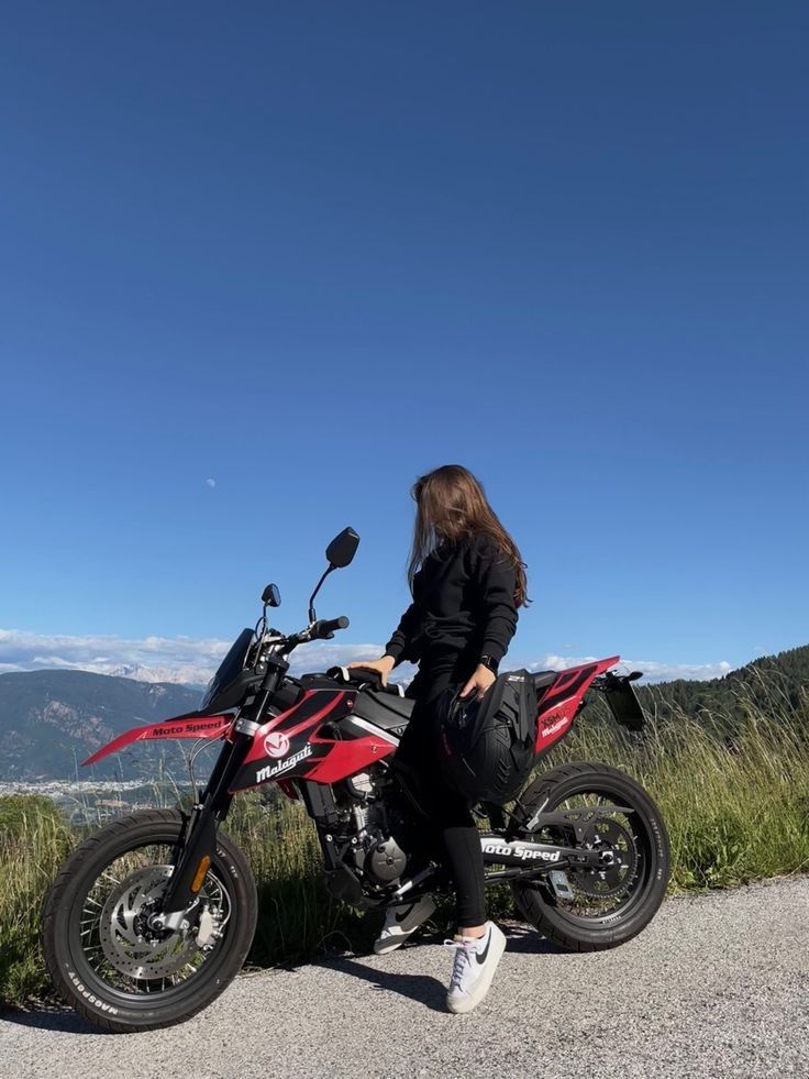 a woman standing next to a red motorcycle on top of a dirt road with mountains in the background