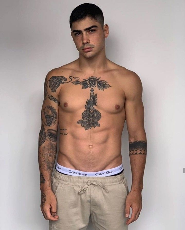 a man with tattoos on his chest standing in front of a white wall wearing khaki shorts