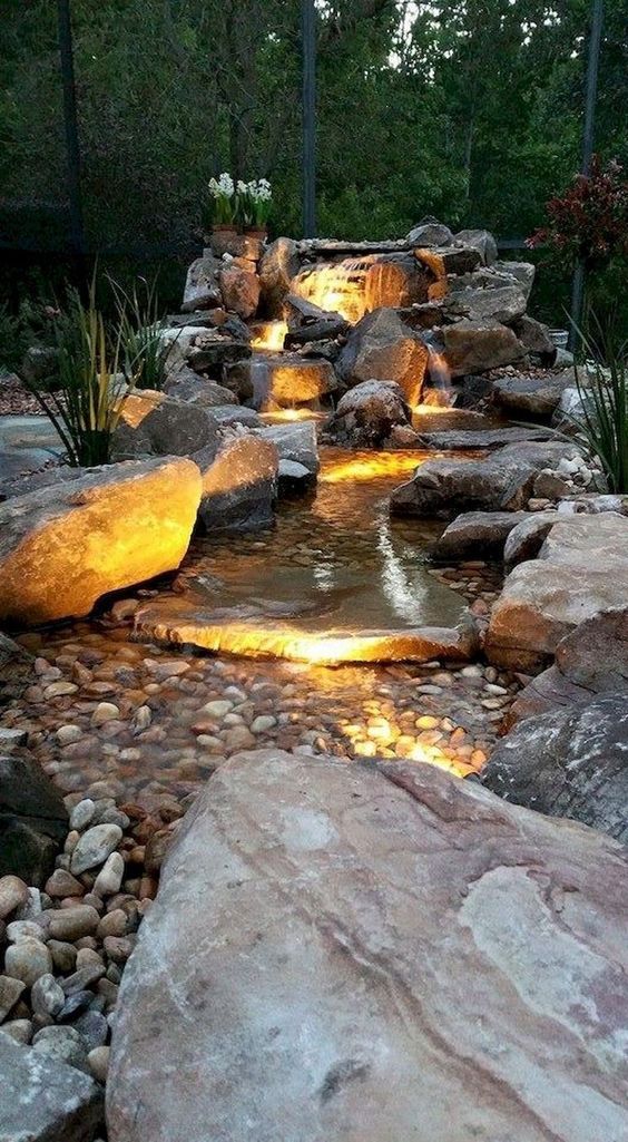 an outdoor pond surrounded by rocks and water features lighting up the area at night time