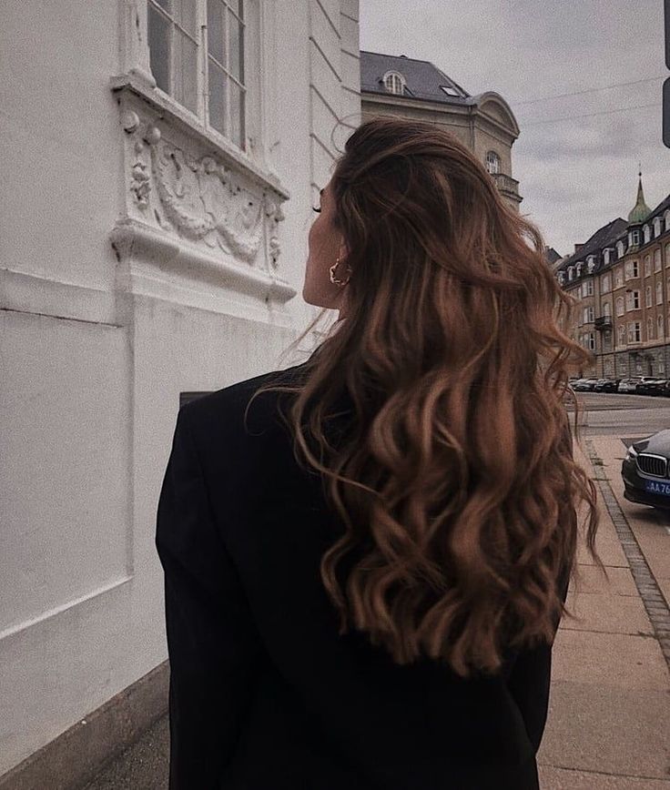 a woman with long, wavy hair standing on the side of a street in front of a building