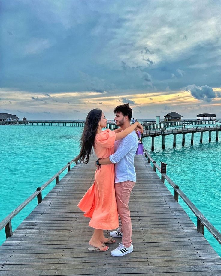 a man and woman standing on a pier near the ocean with their arms around each other