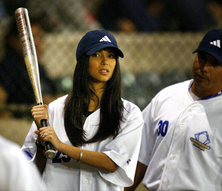 a woman holding a baseball bat while standing next to a man on a field with other people behind her