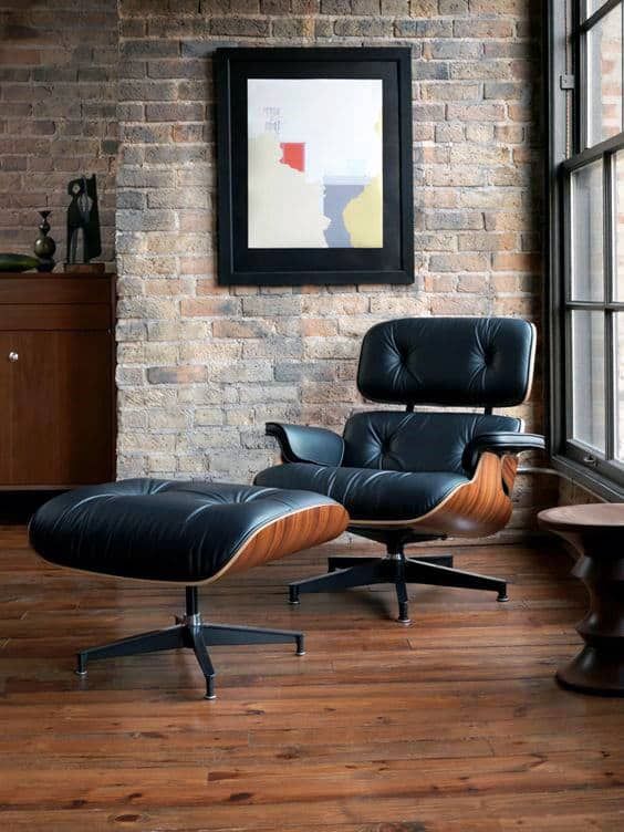 an eames chair and ottoman in front of a brick wall with a painting on it