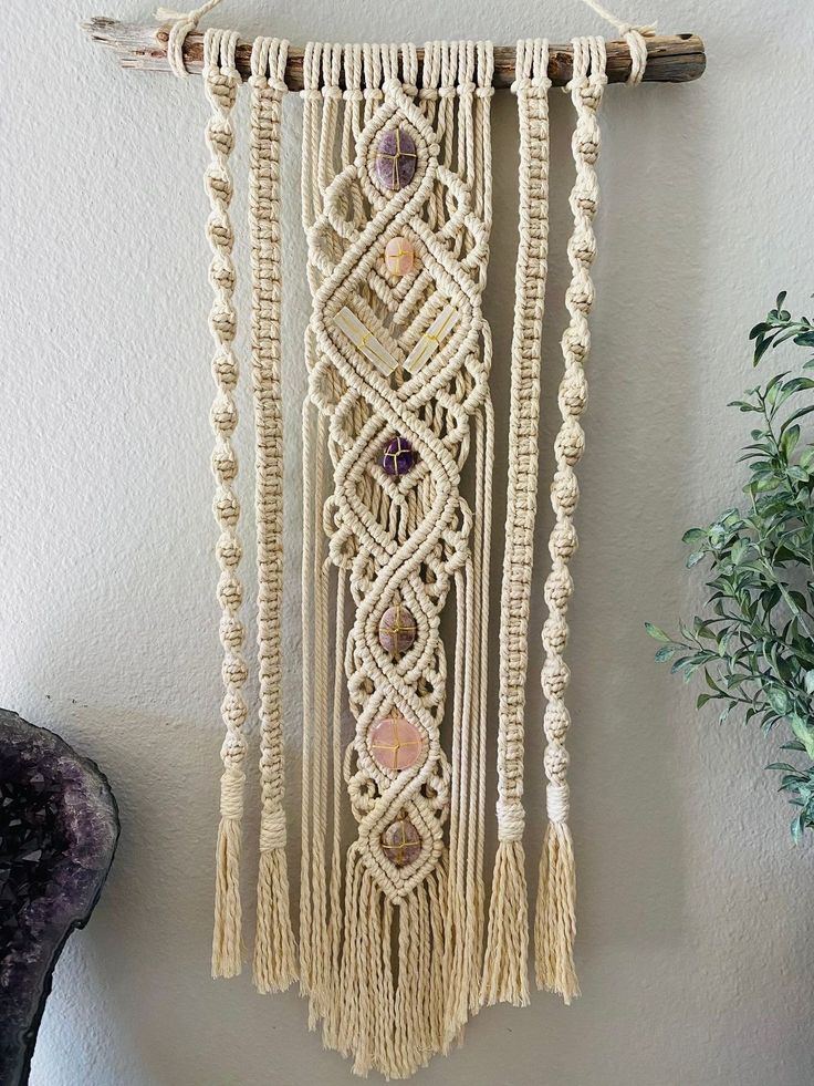 macrame wall hanging with beads and stones on wood sticks, next to potted plant