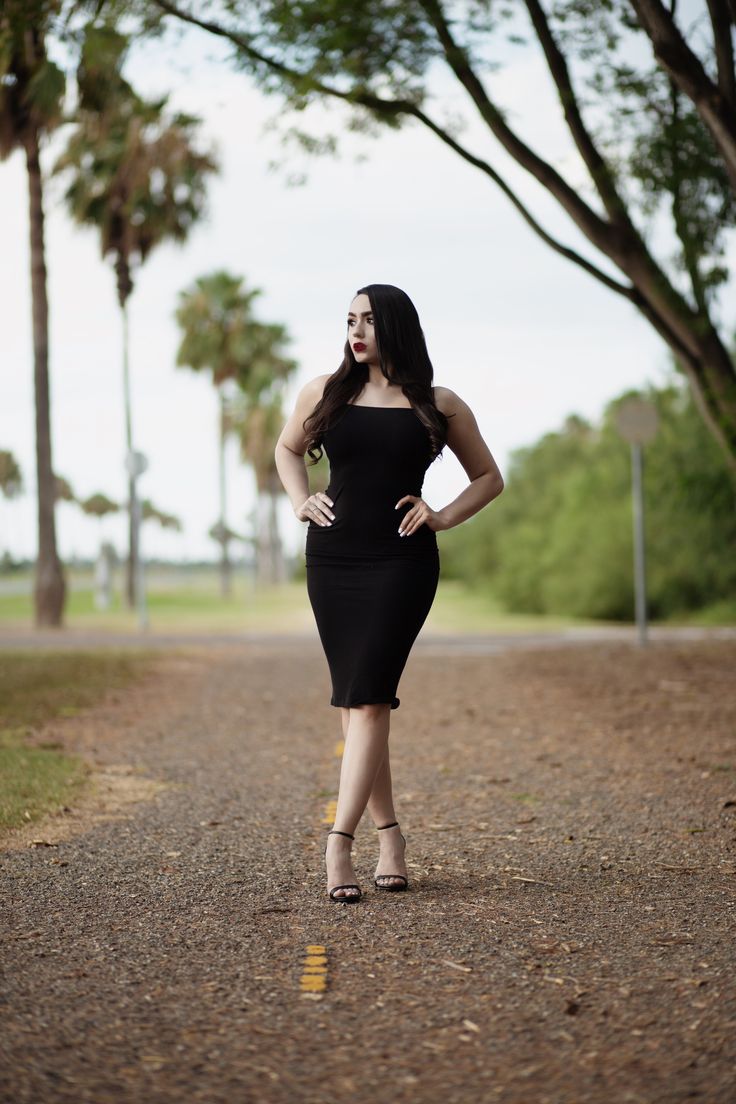 a woman in a black dress is standing on the road with her hands on her hips