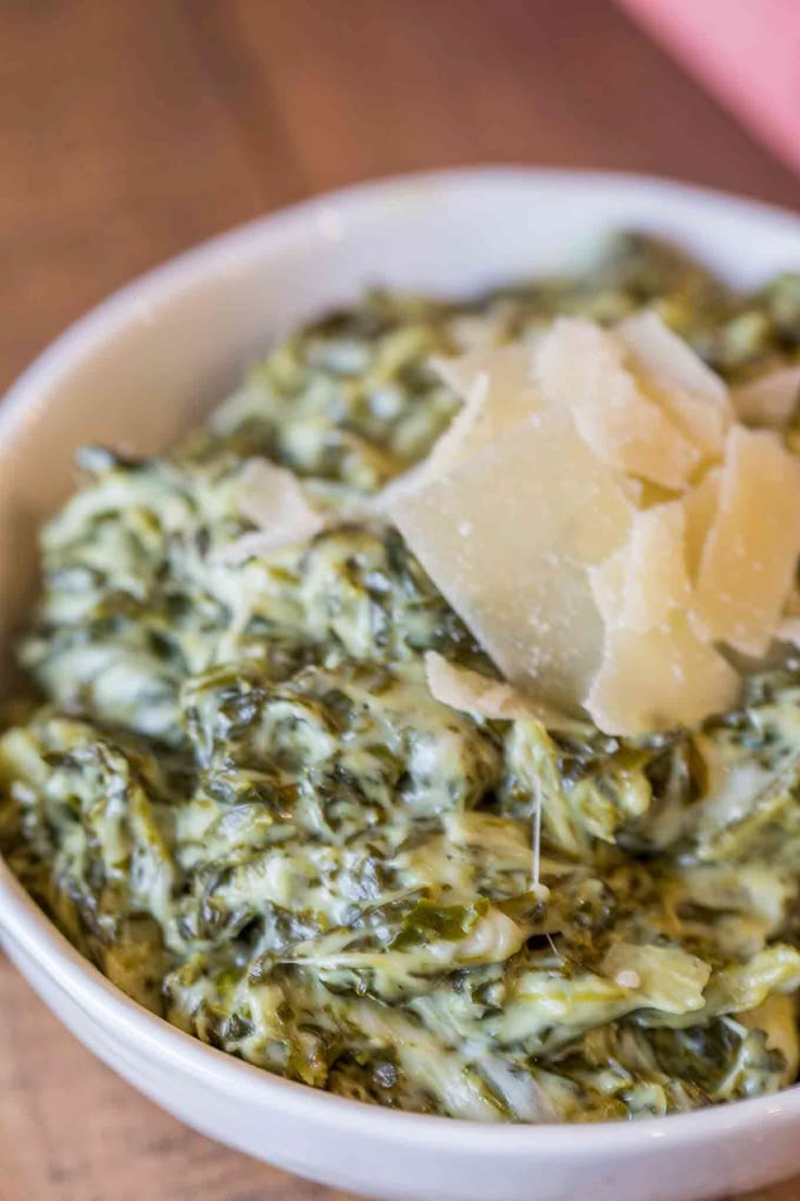 a white bowl filled with spinach and cheese on top of a wooden table next to a pink napkin