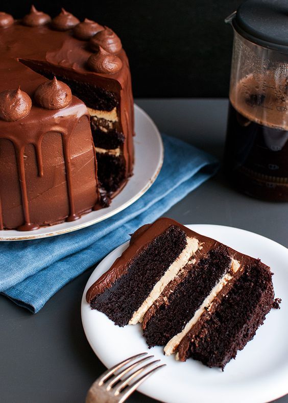 a slice of chocolate cake on a plate next to a coffee pot and fork with the rest of the cake in front of it