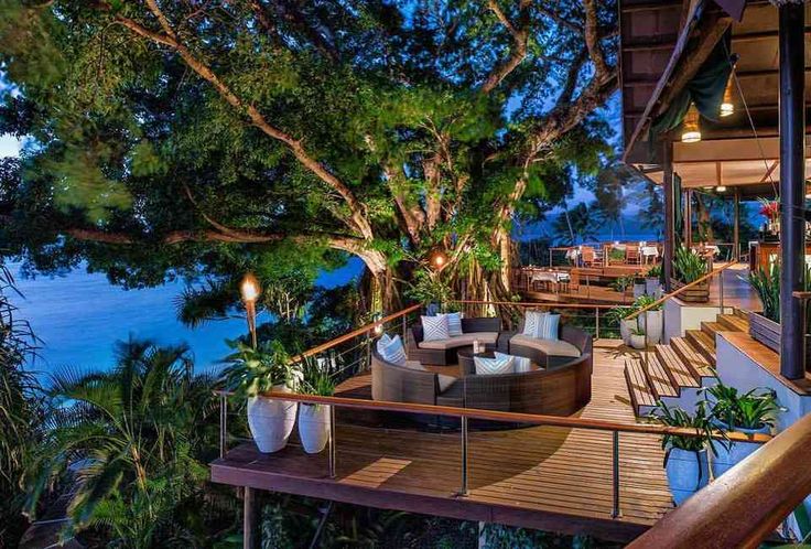 an outdoor deck with chairs and tables next to the ocean at night, surrounded by greenery