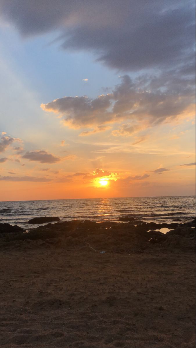 the sun is setting over the ocean with clouds in the sky and some rocks on the beach