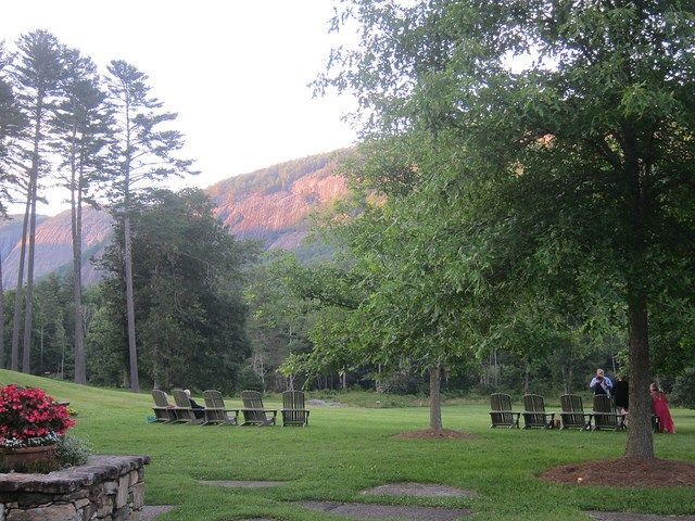 two people are standing in the grass near some trees and lawn chairs, with mountains in the background