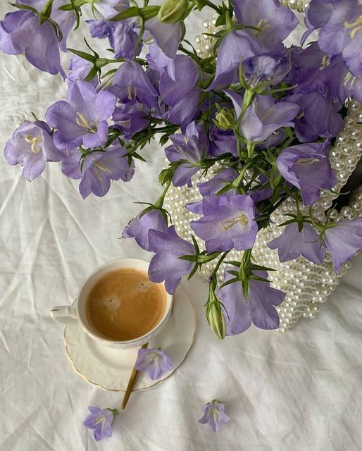 a cup of coffee and some purple flowers on a white table cloth with pearls in the background