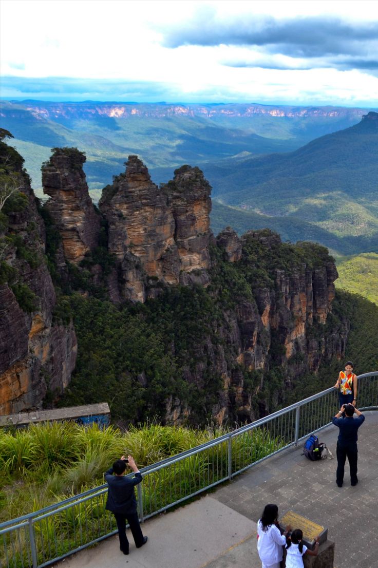 people standing on the edge of a cliff looking out at some mountains and trees in the distance