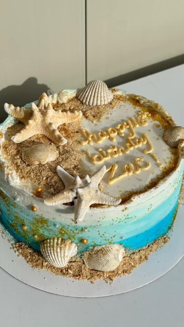 a birthday cake decorated with sea shells and writing on the frosting is sitting on a table