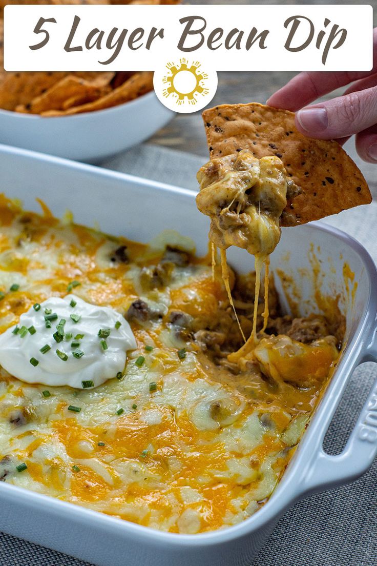 a person dipping a tortilla chip into a casserole dish with cheese and sour cream