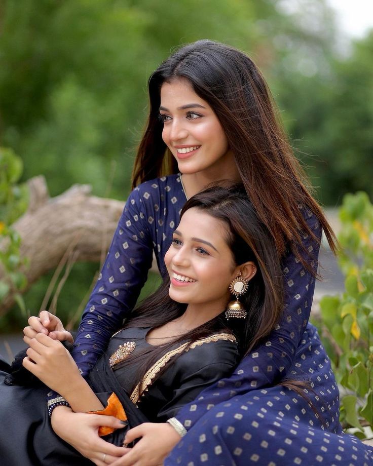 two young women sitting next to each other in front of trees and bushes, both smiling at the camera