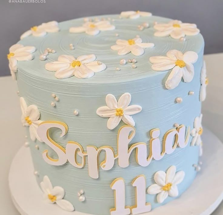 a blue and white birthday cake with daisies on the top is decorated with gold lettering