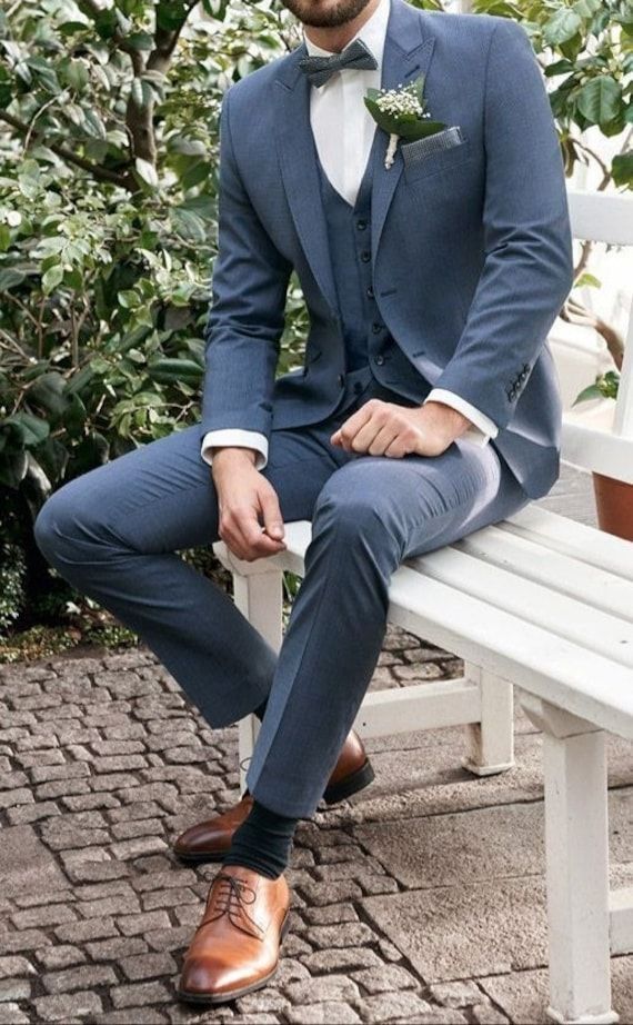 a man in a blue suit sitting on a white bench wearing brown shoes and a boutonniere