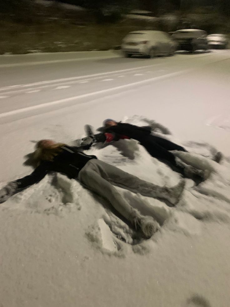 two people laying in the snow next to each other on a road with cars behind them